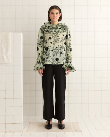 In a tiled room, someone stands expressionless, wearing black pants and the Flocked Sweetunia Top, a handmade, floral blouse inspired by the 1970s with ruffles.