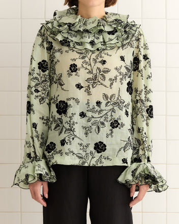 The person stands against a tiled wall, wearing the Flocked Sweetunia Top—a light green floral 1970s blouse with tiered ruffles, featuring a ruffled collar and cuffs.