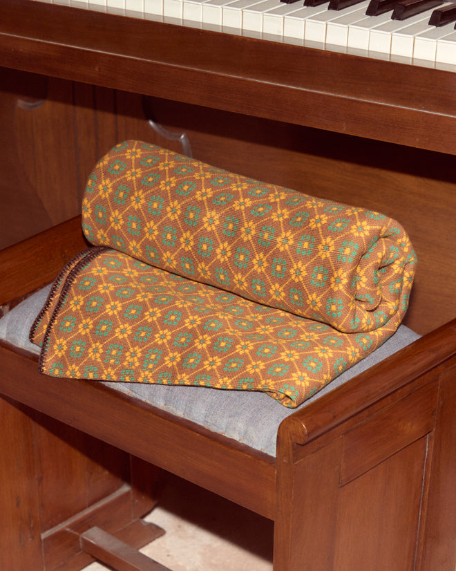 The Floral Jacquard Throw - Brown is rolled up and placed on a bench beneath a wooden piano keyboard.