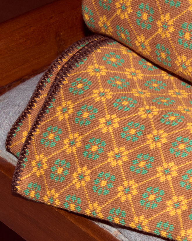 The Floral Jacquard Throw - Brown, a patterned cotton blanket with yellow and green accents made in Peru, is draped over a wooden bench with a gray cushion.