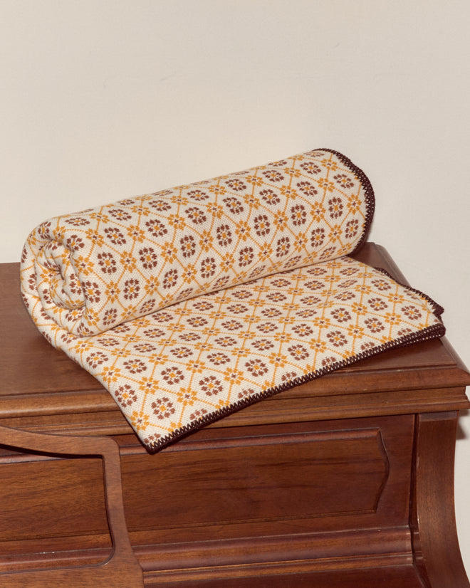 The Floral Jacquard Throw - Marigold, with its vintage yellow and brown pattern, is rolled and displayed on a wooden bench, bringing charm to your home decor collection.