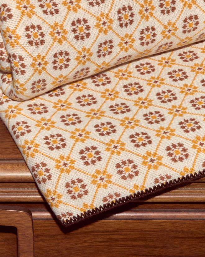 The Floral Jacquard Throw - Marigold, a beige cotton throw with a yellow and brown floral diamond pattern, rests folded on a wooden surface—ideal for bringing vintage charm to your home décor collection.