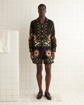 A man stands in a tiled room wearing a matching black 1960s-inspired outfit adorned with colorful floral and geometric patterns. He is dressed in an embroidered Floral Pinwheel Long Sleeve Shirt paired with 100% linen shorts and black shoes.