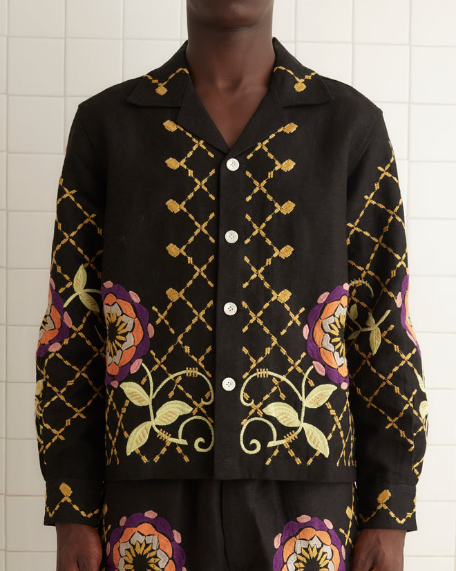 A man stands in a tiled room wearing a matching black 1960s-inspired outfit adorned with colorful floral and geometric patterns. He is dressed in an embroidered Floral Pinwheel Long Sleeve Shirt paired with 100% linen shorts and black shoes.