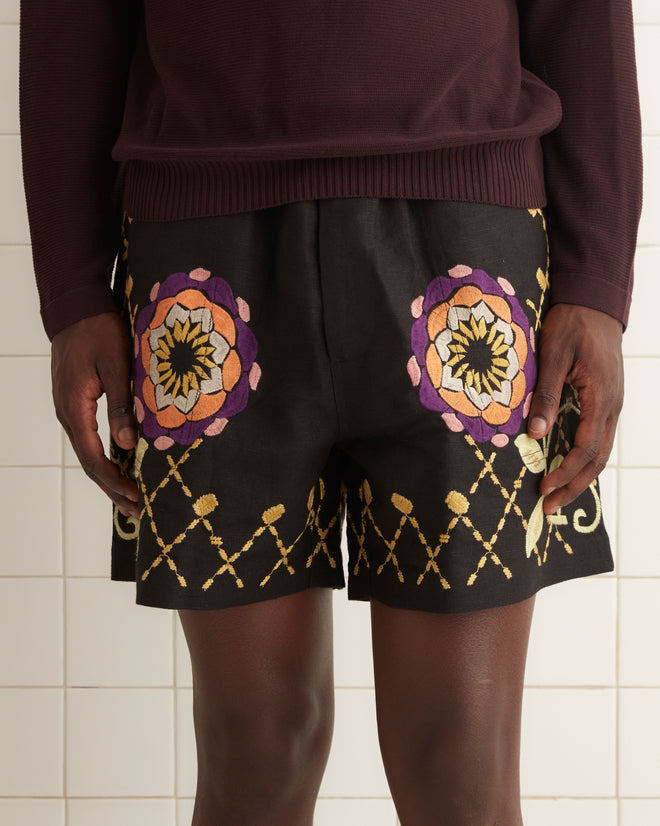 A person is standing in a white-tiled room, wearing a dark long-sleeve shirt and black shoes. They have on the Floral Pinwheel Shorts, which feature a colorful satin-stitched pattern, lending a vibrant touch to their outfit as they appear to be looking straight ahead.