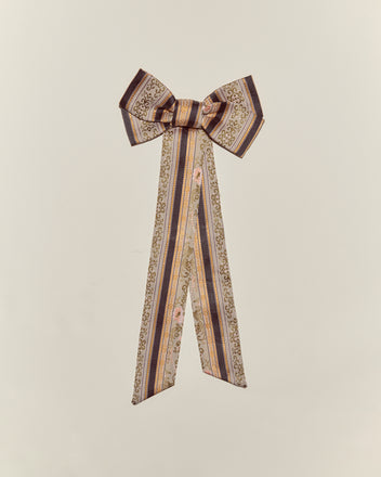 The Floret Brocade Bow Barrette features long, patterned ribbons in gold, brown, and pink, creating a decorative handmade accessory on a plain background.