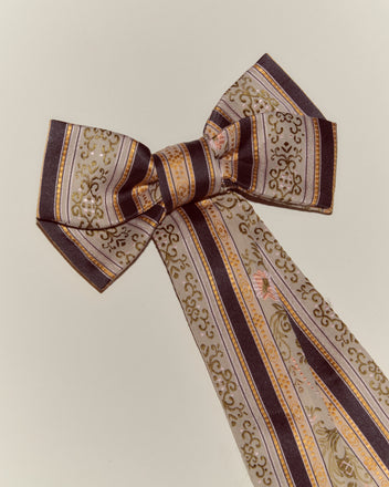 The Floret Brocade Bow Barrette features brown, gold, and lavender stripes with ornate patterns, capturing the look of a floral brocade bow on a light background.