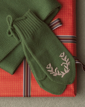 The Floret Mittens in green, featuring beautiful hand-embroidered florals, are elegantly placed on a red, ribbon-wrapped gift box next to a matching green scarf.