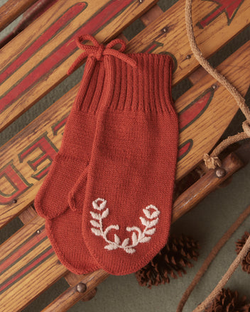 The Floret Mittens - Red, crafted from soft merino wool and adorned with intricate white floral embroidery, are displayed on a wooden sled, encircled by pinecones and rope.