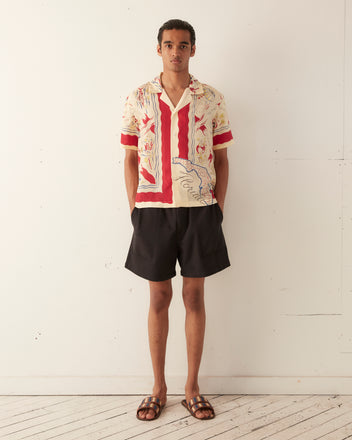 A person stands against a plain wall wearing the Florida Map Short Sleeve Shirt made of 100% silk, accompanied by black shorts and sandals.