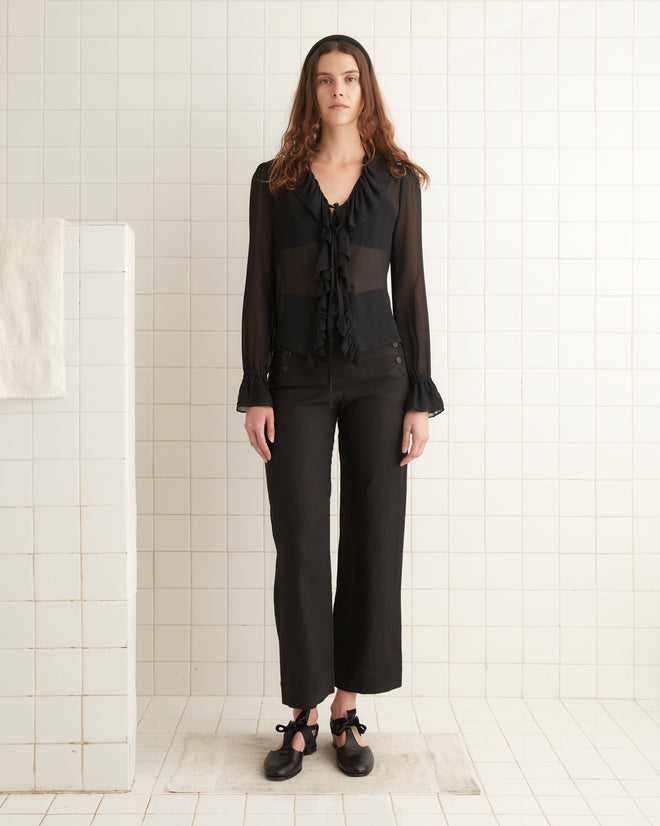 A woman stands in a tiled room, wearing a Flounce Blouse - Black paired with black pants. She has long, wavy hair and faces the camera directly. The room features a white towel and a rectangular block structure, contrasting with her lightweight sheer silk ensemble.