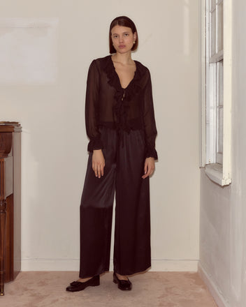 A woman wearing the Flounce Blouse - Black stands indoors by a window, looking at the camera.