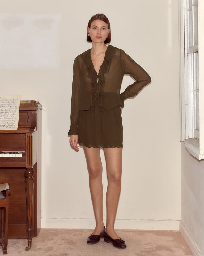 Woman in a sheer dark outfit featuring the Flounce Blouse in olive stands by a piano with sheet music, in a bright room with a window and carpeted floor.