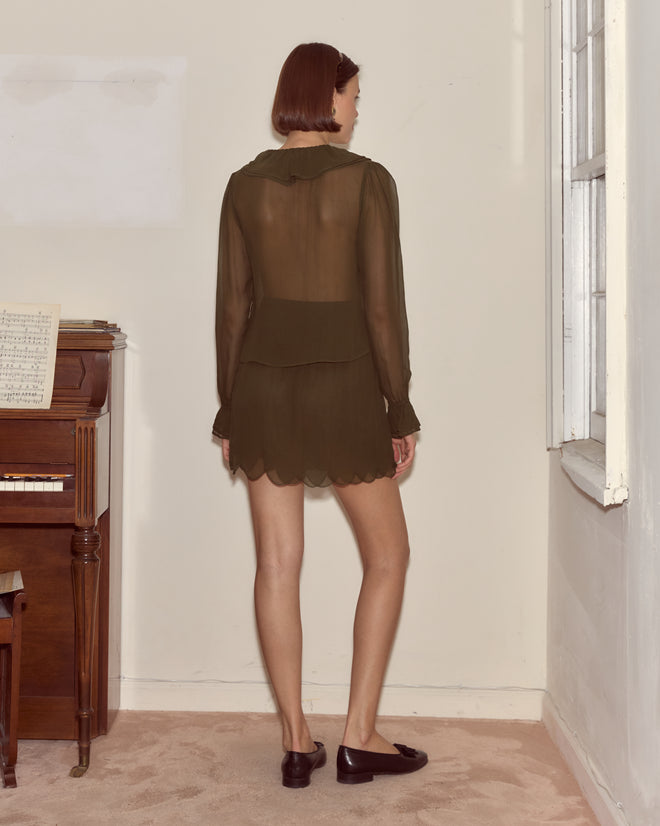 A woman in the Flounce Blouse - Olive stands by a piano and window facing a wall, her sheer top echoing a timeless French silhouette in the carpeted room.