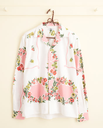 The Flowered Terrace Shirt (XS/S), a unique white long-sleeve buttoned shirt reminiscent of a midcentury tablecloth, features a vibrant pink, green, and red floral pattern and hangs on a hook against a cream-colored wall.