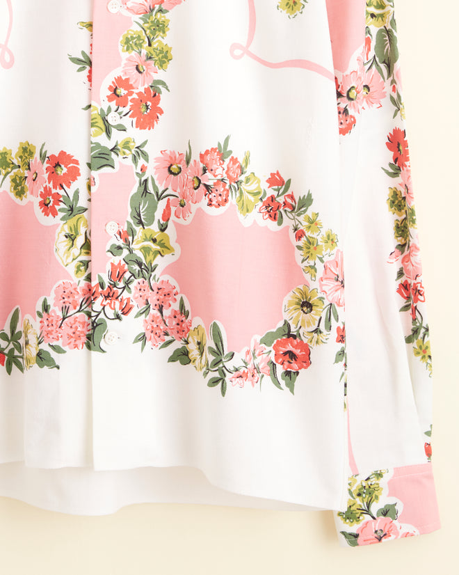 The Flowered Terrace Shirt (XS/S), a unique white long-sleeve buttoned shirt reminiscent of a midcentury tablecloth, features a vibrant pink, green, and red floral pattern and hangs on a hook against a cream-colored wall.