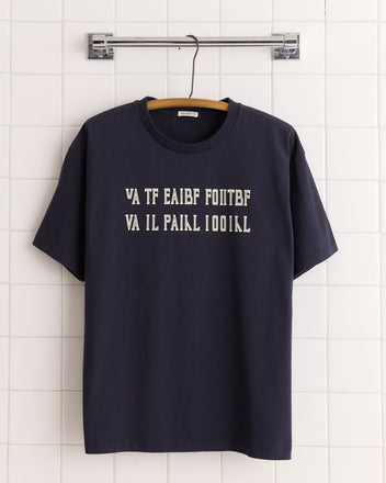 The Folding Puzzle Tee, a black 100% cotton shirt with white code-style text, hangs on a wooden hanger against tiled walls, suggesting it contains a hidden message.