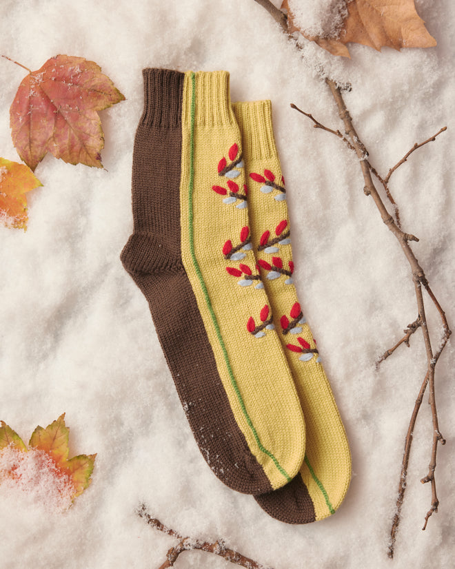 Amidst the snowy setting, the Foliage Socks—a pair of cozy accessories crafted from soft merino wool—stand out with one sock in brown and the other adorned with yellow and red floral patterns. Scattered leaves and twigs surround them, enhancing their charm against the wintry backdrop.