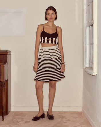 A woman stands indoors wearing the Folly Bra, a black burlesque bralette, paired with a black-and-white striped skirt and black flats.
