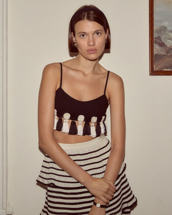 Woman indoors near a framed painting wears the Folly Bra, a burlesque-style bralette with tassels, paired with a striped skirt.