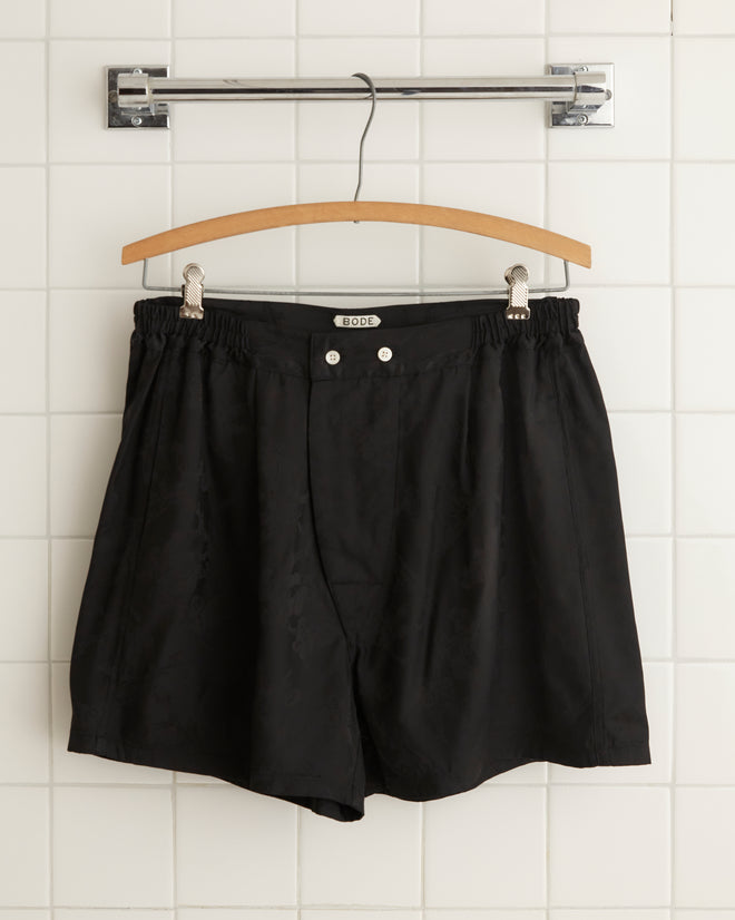 The Football Jacquard Boxers in black, featuring an elastic waistband and buttons, are hanging from a wooden hanger on a metal rod against a tiled wall background.