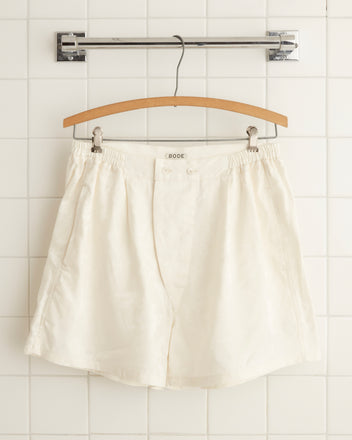 Cream Football Jacquard Boxers hanging on a wooden hanger against a tiled wall. The design includes a subtle football pattern and the waistband features two front buttons with an elasticized back. A 