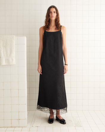 A woman stands in a tiled bathroom, wearing the Football Jacquard Column Gown with a classic column silhouette and black flats.