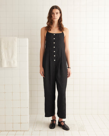 A woman is shown from the back wearing a black Football Jacquard Jumpsuit with crisscross straps and a bow tied at the lower back. She stands in front of a tiled wall, her silhouette suggesting the elegance of a midcentury-inspired design.