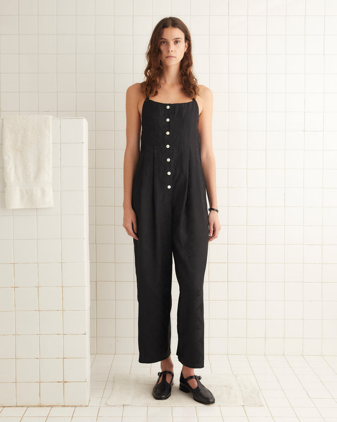 A woman is shown from the back wearing a black Football Jacquard Jumpsuit with crisscross straps and a bow tied at the lower back. She stands in front of a tiled wall, her silhouette suggesting the elegance of a midcentury-inspired design.