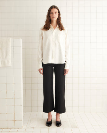 A person stands in a minimalist white-tiled room wearing the Football Jacquard Murray Shirt with its subtle woven pattern, black pants, and black shoes.