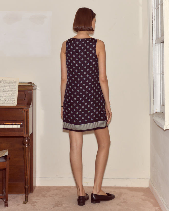 A woman in the Foyer Diamond Dress, featuring a sleeveless Mod silhouette and 1930s cravat print, stands with her back to the camera by a piano in a sunlit room.