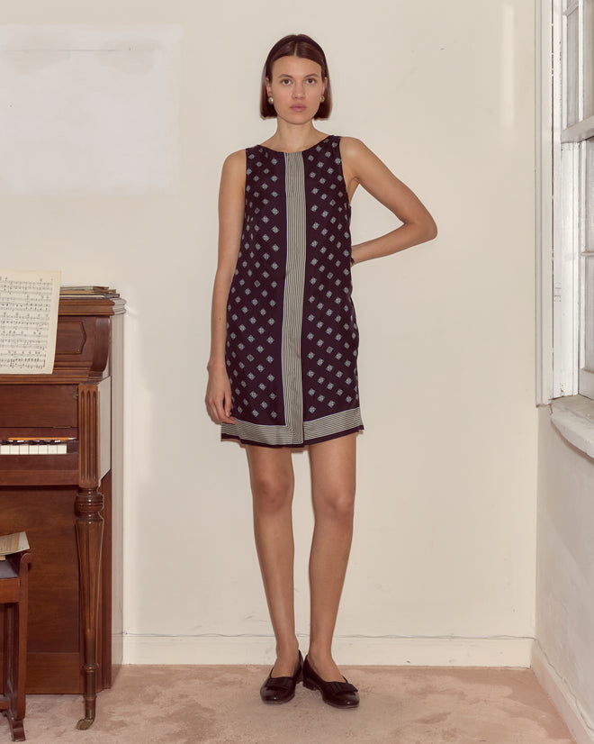 A woman in the Foyer Diamond Dress, featuring a Mod silhouette and 1930s cravat print, stands indoors by a piano and window with one arm bent at her waist.