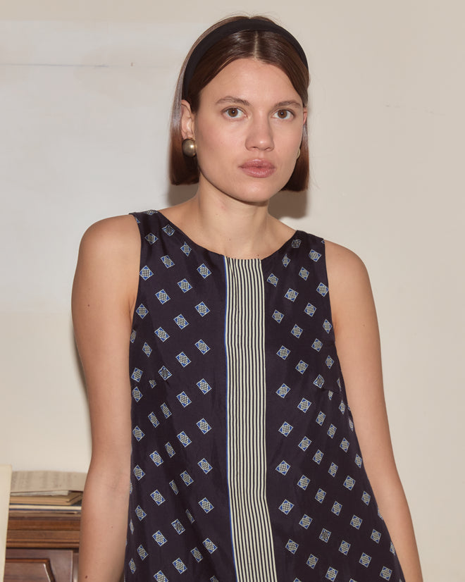 Woman with straight brown hair, wearing the Foyer Diamond Dress—navy with a subtle 1930s cravat print—and pearl earrings, faces camera.