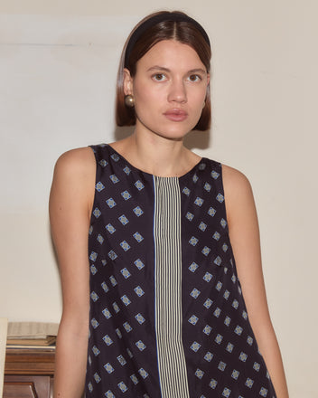 Woman with straight brown hair, wearing the Foyer Diamond Dress—navy with a subtle 1930s cravat print—and pearl earrings, faces camera.