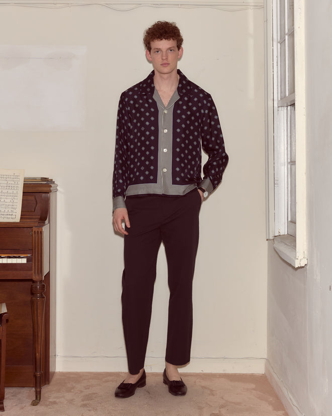 A man stands indoors by a piano, wearing the Foyer Diamond Long Sleeve Shirt, black trousers, and loafers.