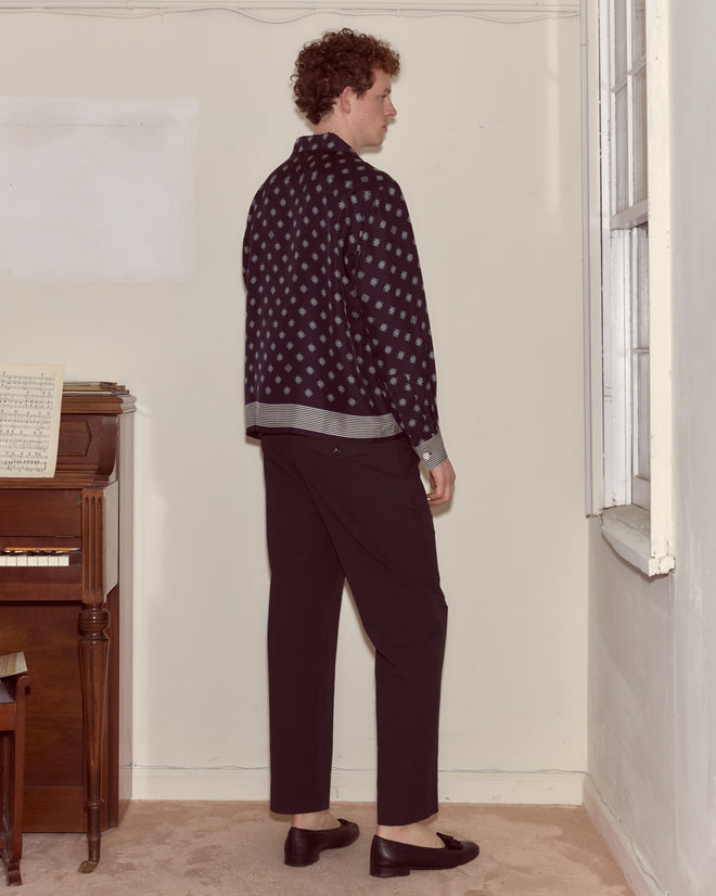 A man in a Foyer Diamond Long Sleeve Shirt stands facing a corner beside a piano and a window in a sparsely furnished room.