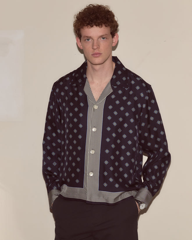 Young man with curly hair wears the Foyer Diamond Long Sleeve Shirt, a patterned navy silk foulard with white accents, standing against a beige wall.
