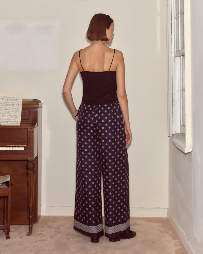 A woman in a black top and Foyer Diamond Pants stands facing a wall near a piano and window.