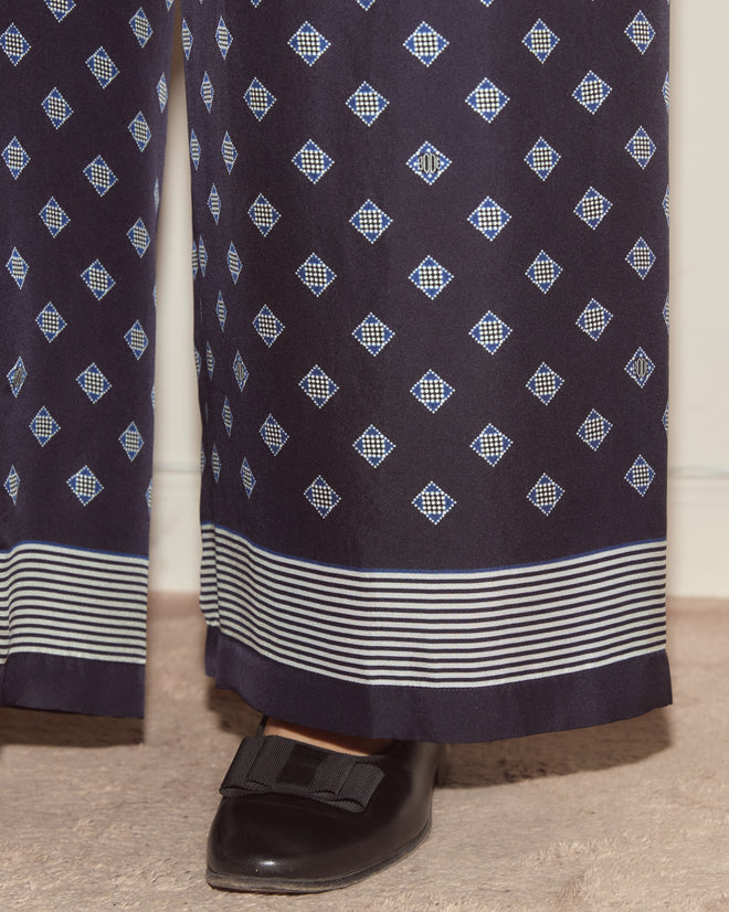 The Foyer Diamond Pants showcase a navy foulard print with a diamond pattern and striped hem, styled with black shoes adorned with a bow detail.