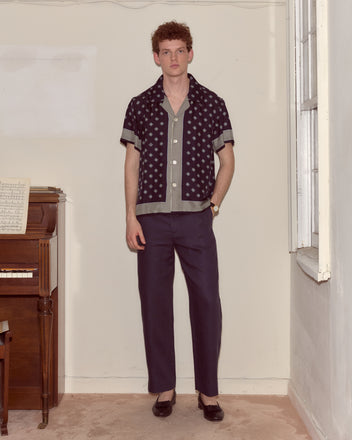 A man in a Foyer Diamond Short Sleeve Shirt and dark pants stands by a piano in a simple, sunlit room.
