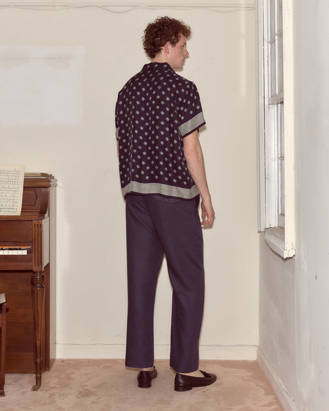 A man in a Foyer Diamond Short Sleeve Shirt stands facing a corner by a piano and a window in a sparsely furnished room.