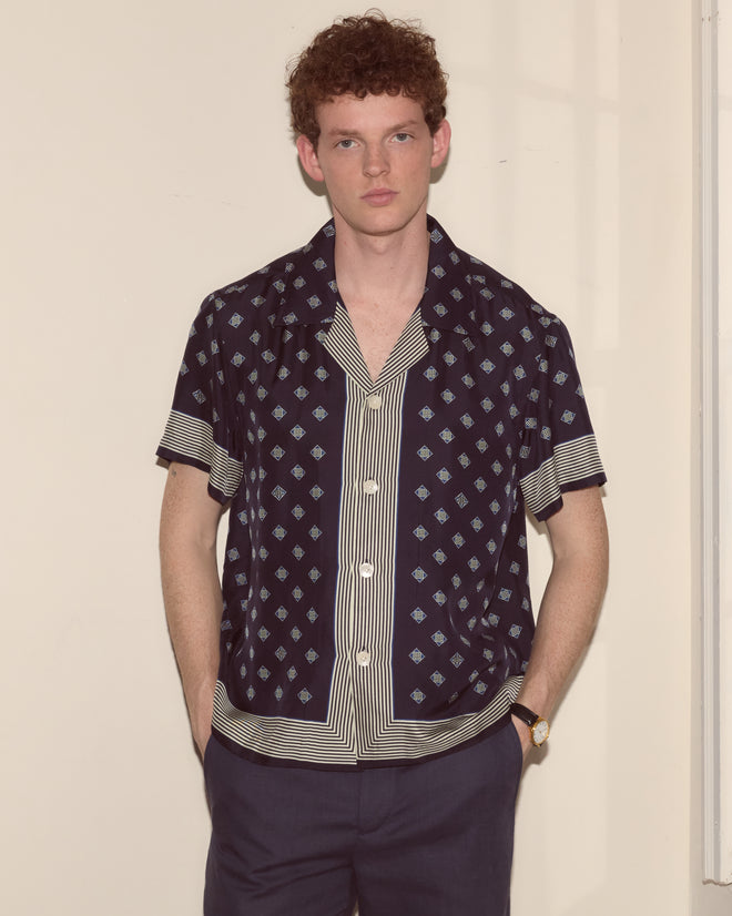A man with curly hair wears the Foyer Diamond Short Sleeve Shirt and dark pants, standing with his hands in his pockets.
