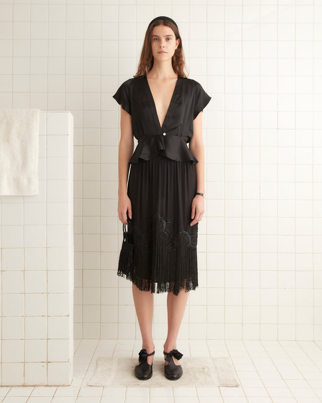 A woman stands in a tiled room wearing the black Freya Top paired with a pleated skirt and black silk shoes. A white towel hangs on a bar to the left.