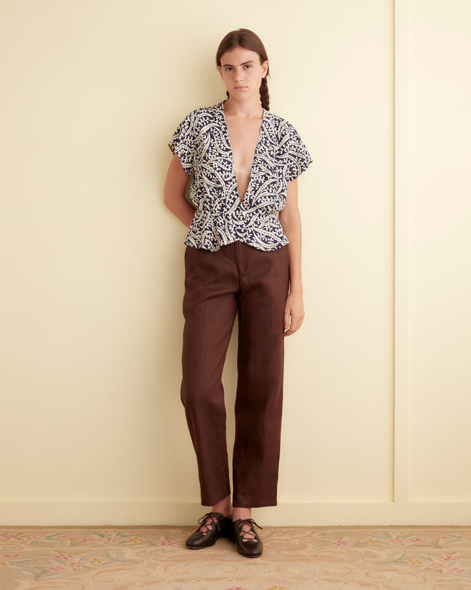 A woman wears the Friendly Fern Top—a unique deadstock rayon piece with a navy fern print—paired with brown pants and dark shoes, standing by a pale wall.