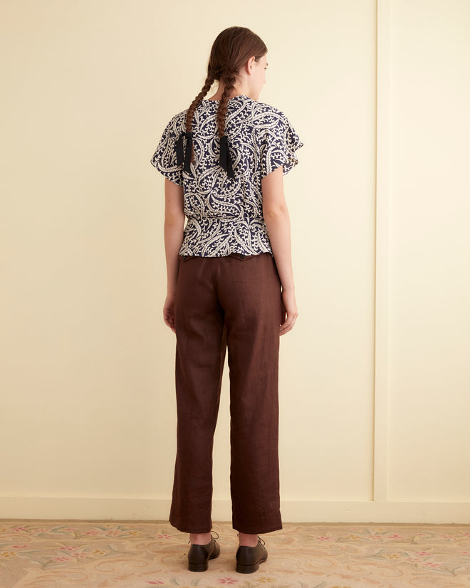 A person with braided hair, wearing the Friendly Fern Top crafted from unique deadstock fabric and brown pants, stands in a room facing a light-colored wall.