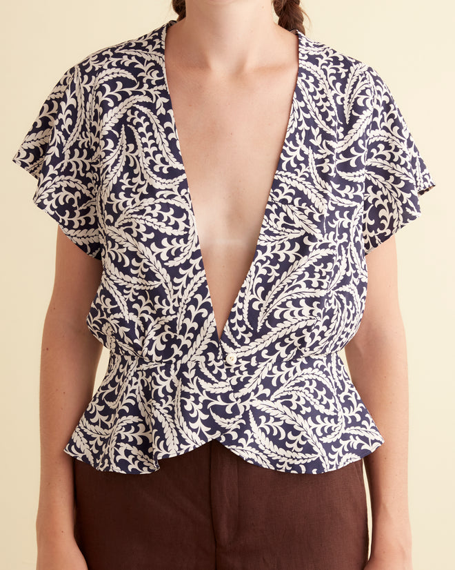 A woman stands against a beige background, wearing the one-of-a-kind Friendly Fern Top—a deep V-neck deadstock rayon shirt with a navy fern pattern and short sleeves—paired with brown pants.