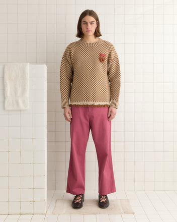 A person with long hair in a Fringe Check Pullover - Brown and pink pants stands in a tiled room near a white towel draped over a wall.