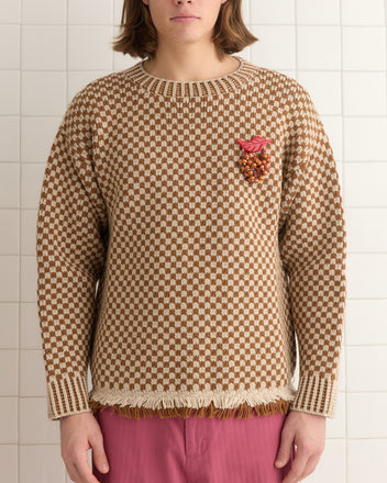 A person wearing a Fair Trade Certified Fringe Check Pullover in brown, crafted from merino wool and featuring a small red and orange embellishment, paired with stylish pink pants.