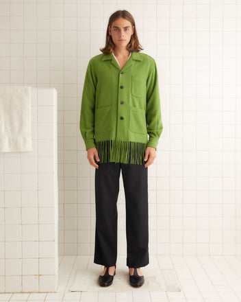 In a tiled room, a person wears the Fringe Overshirt, a green jacket with midcentury flair, paired seamlessly with black pants and shoes.