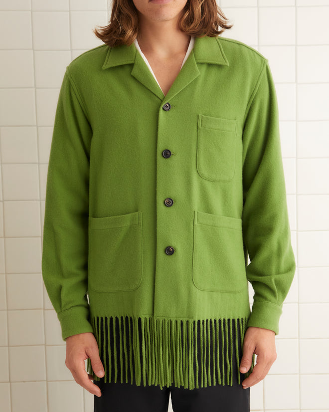 In a tiled room, a person wears the Fringe Overshirt, a green jacket with midcentury flair, paired seamlessly with black pants and shoes.